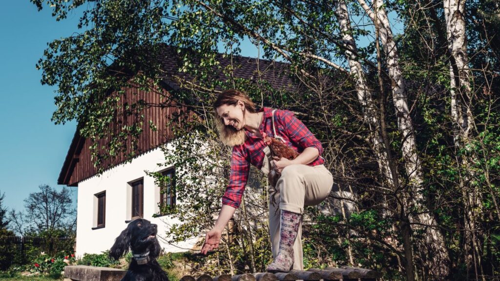 Žena v károvanej košeli so psom na trávnatej záhrade pred bielou chalupou s dreveným štítom a vysokými brezami.