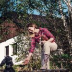 Žena v károvanej košeli so psom na trávnatej záhrade pred bielou chalupou s dreveným štítom a vysokými brezami.