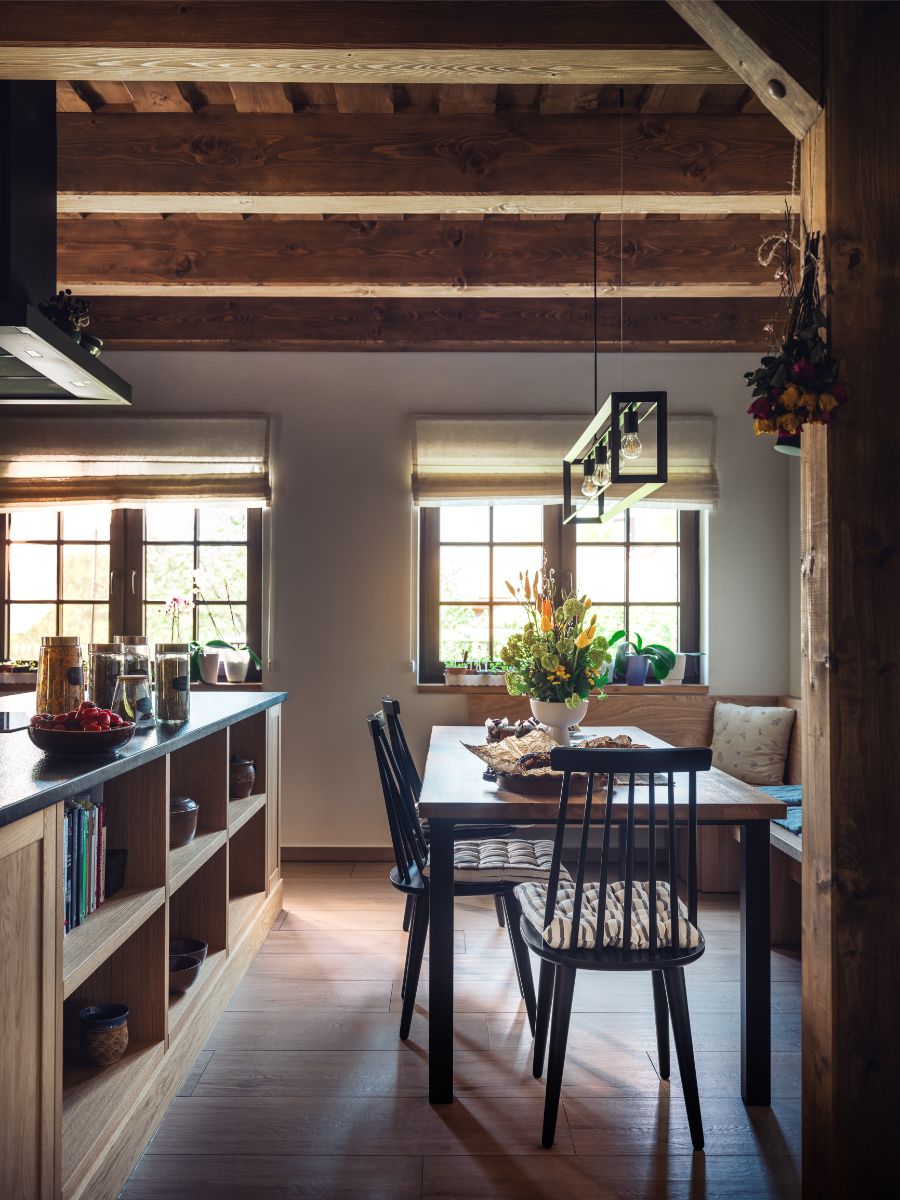 Drevený jedálenský stôl s čiernymi stoličkami pri oknách s béžovými rímskymi roletami v chalupárskom štýle.