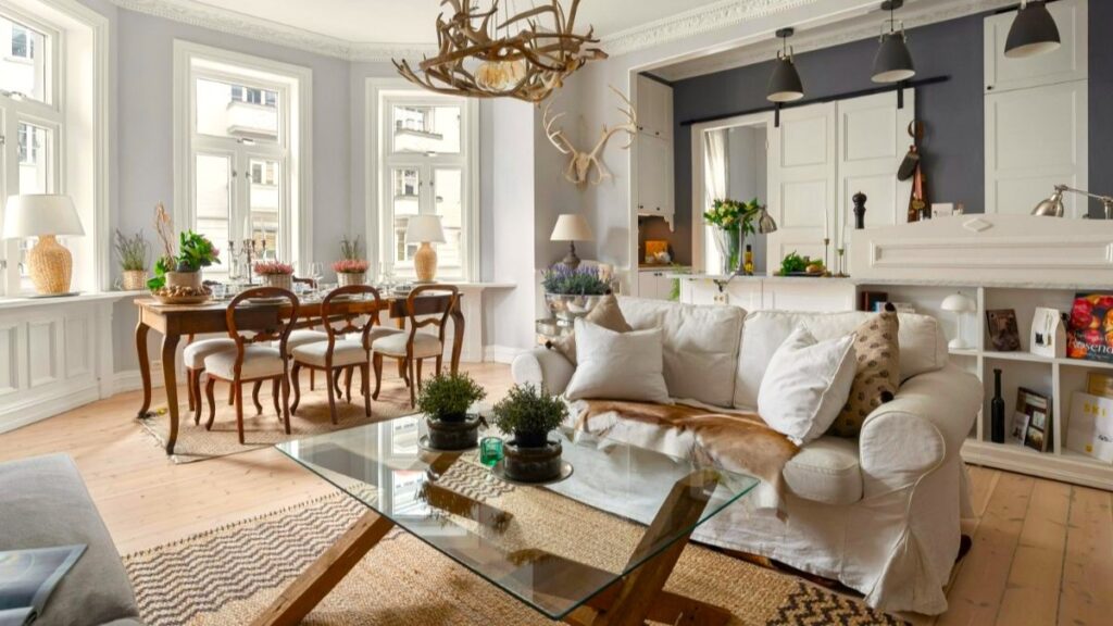 Biely moderný interiér s rustikálnym parožinovým lustrom, historickým dreveným stolom a svetlou textilnou sedačkou.