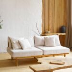 Minimalistická biela pohovka na drevených nohách s organickým konferenčným stolíkom a hlineným závesným svietidlom.