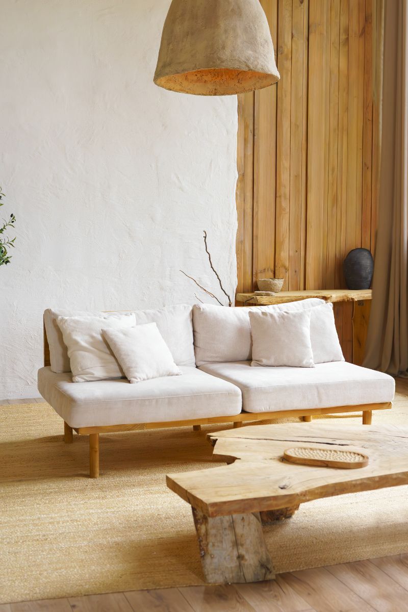 Minimalistická biela pohovka na drevených nohách s organickým konferenčným stolíkom a hlineným závesným svietidlom.