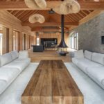 Otvorený drevený krov nad visiacimi prútenými lustrami, čierny závesný krb a masívny dubový stôl v obývacej izbe.