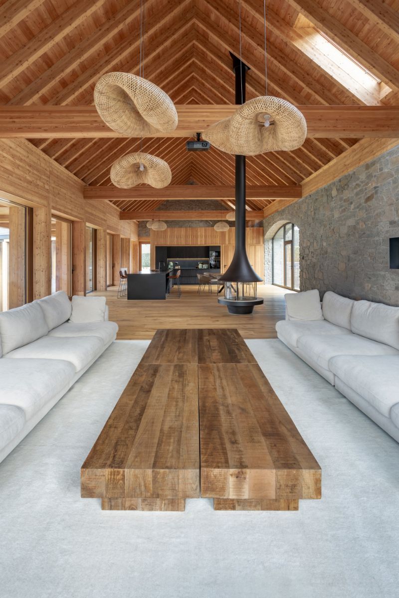 Otvorený drevený krov nad visiacimi prútenými lustrami, čierny závesný krb a masívny dubový stôl v obývacej izbe.