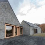 Sivá kamenná fasáda s vertikálnym dreveným obkladom, svetlé smrekovcové rámy okien a tmavá šikmá plechová strecha.