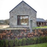 Štítová stena z prírodného kameňa s veľkým oknom, nízky kamenný plot so stĺpikom a okrasná zeleň.