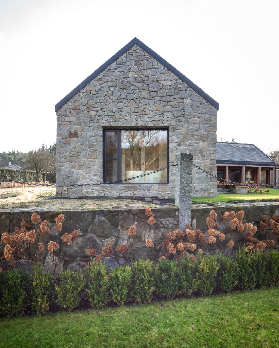 Štítová stena z prírodného kameňa s veľkým oknom, nízky kamenný plot so stĺpikom a okrasná zeleň.