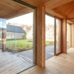 Presklená drevená stena pri svetlej dubovej podlahe a vonkajšia terasa s výhľadom na kamennú stodolu.