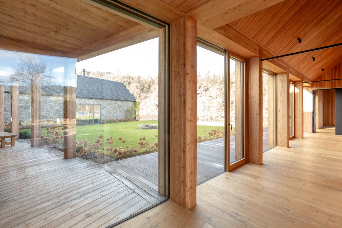 Presklená drevená stena pri svetlej dubovej podlahe a vonkajšia terasa s výhľadom na kamennú stodolu.
