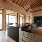 Čierny kuchynský ostrov s koženými barovými stoličkami pred presklenou stenou s masívnym dreveným rámom.