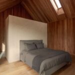 Sivá posteľ pred béžovou deliacou stenou pod šikmým dreveným stropom so zapusteným strešným oknom.
