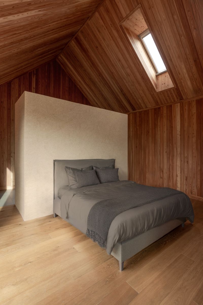 Sivá posteľ pred béžovou deliacou stenou pod šikmým dreveným stropom so zapusteným strešným oknom.