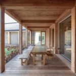 Masívny drevený stôl s lavicou pod trámovým stropom terasy a výhľad na kamennú fasádu vedľajšej budovy.