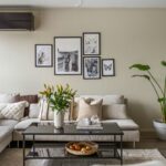 Béžová sedačka a čierny kovový stolík s dekoráciami a vázou s kvetmi pod nástennou galériou na svetlom huňatom koberci.