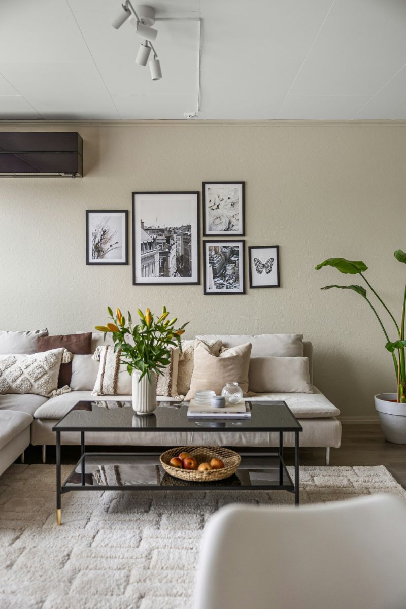 Béžová sedačka a čierny kovový stolík s dekoráciami a vázou s kvetmi pod nástennou galériou na svetlom huňatom koberci.