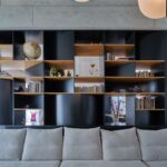 Čierna asymetrická knižnica s drevenými policami za svetlosivou látkovou pohovkou v modernom interiéri.