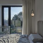 Moderná spálňa s posteľou so sivým čalúneným čelom postele, detskou postieľkou a čiernym závesným svietidlom.