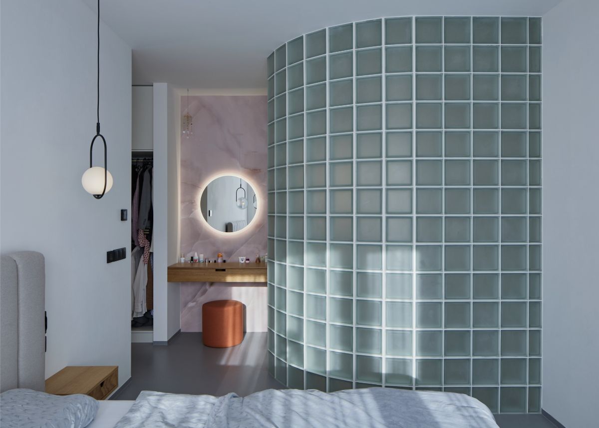 blá stena z matných sklobetónových tvárnic v spálni s ružovou mramorovou stenou a podsvieteným okrúhlym zrkadlom.