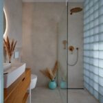 Otvorený sprchový kút s mosadznou batériou, sklobetónovou stenou, bielym umývadlom a béžovou stierkou.