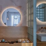 Otvorený drevený organizér na šperky v kúpeľni s podsvieteným zrkadlom a bielym keramickým umývadlom na doske.