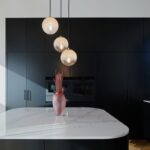 ri guľaté sklenené svietidlá nad bielym kamenným ostrovom s ružovou vázou pred čiernou nábytkovou stenou.