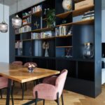 Ružové čalúnené kreslo pri drevenom jedálenskom stole pred čiernou asymetrickou knižnicou s dekoráciami.