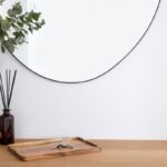 Okrúhle zrkadlo s čiernym rámom, drevený podnos s kľúčmi a biela keramická váza na dubovej komode.