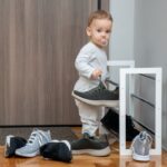 Dieťa v sivom oblečení držiace tmavú športovú topánku pri modernom bielom stojane na obuv.