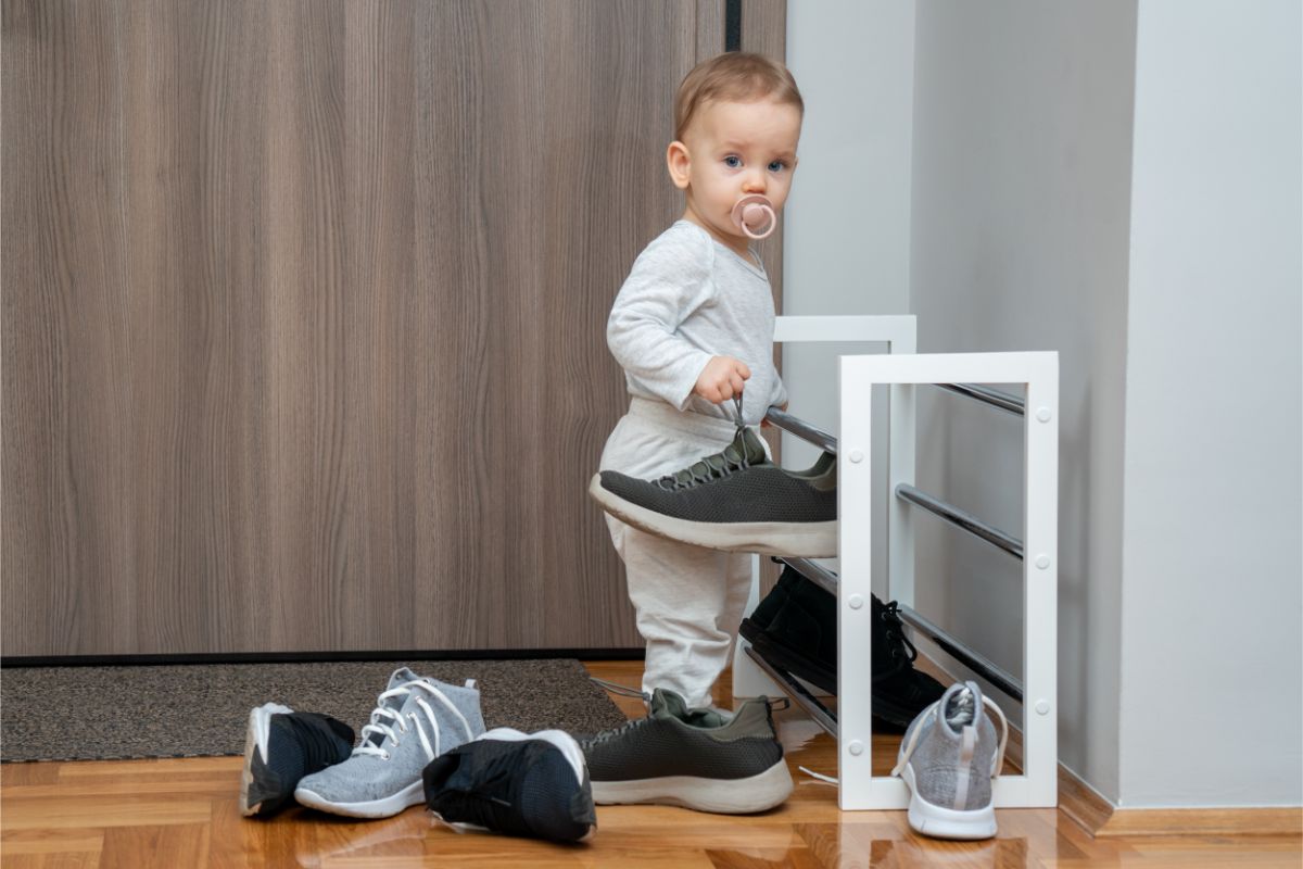 Dieťa v sivom oblečení držiace tmavú športovú topánku pri modernom bielom stojane na obuv.