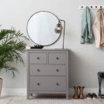 Sivá komoda s okrúhlym zrkadlom a lampou vedľa vešiakov na stene a rastliny na zemi.
