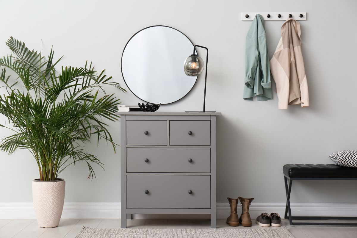 Sivá komoda s okrúhlym zrkadlom a lampou vedľa vešiakov na stene a rastliny na zemi.