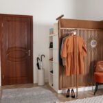 Drevený lamelový panel s vešiakmi, hnedé šaty na ramienku a oranžový zamatový taburet v svetlej chodbe.