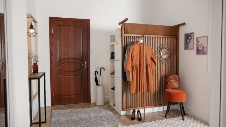 Drevený lamelový panel s vešiakmi, hnedé šaty na ramienku a oranžový zamatový taburet v svetlej chodbe.