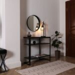 Čierny kovový konzolový stolík s industriálnou lampou a malým okrúhlym zrkadlom na bielej stene.