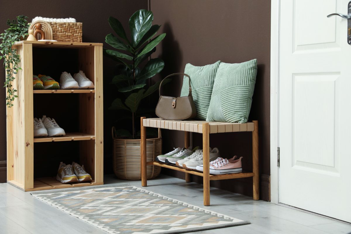 Drevený botník s bielymi teniskami a nízka pletená lavica s modrými vankúšmi pri hnedej stene.