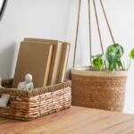 Prútený košík s béžovými knihami a visiaci kvetináč so zelenou rastlinou na drevenom povrchu.