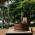 Exteriérová drevená kaďa s oceľovými obručami na drevenej plošine pod zelenými stromami.