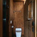 Minimalistická kúpeľňa s bielym závesným WC, vertikálnym dreveným obkladom a obdĺžnikovým zrkadlom.