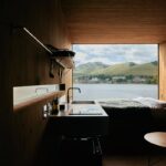 Jednoduchá kuchynská linka, otvorená polica a posteľ na platforme pri veľkom okne v interiéri mobilného domu.