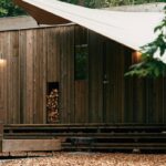 Jedna strana mobilného domu z dreva so schodíkmi, výklenkom na štiepané drevo a tieniacou plachtou.