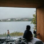 Žena s malým synom, sediaca na posteli pri veľkoformátovom okne s výhľadom na jazero a protiľahlý breh.