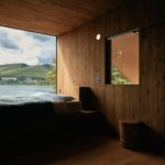 Celodrevený interiér mobilného domu s nízkou posteľou na platforme, malým aj veľkoformátovým oknom a dreveným stolčekom.