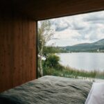 Minimalistická posteľ s olivovozelenou dekou pred veľkoformátovým oknom s výhľadom na jazero.
