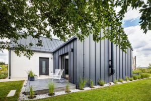 Moderný rodinný dom s plechovou aj bielou fasádou, sedlovou strechou a upravenými záhonmi.