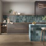 Moderná sivá kuchyňa s modrým obkladom, ostrovček z tyrkysových dlaždíc a svetlá drevená podlaha s barovou stoličkou.