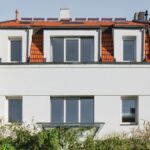 Dom s bielou fasádou, červenou škridlovou strechou, svetlým plným plotom a bujnou zeleňou.