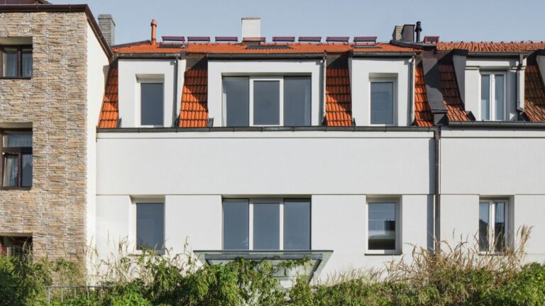 Dom s bielou fasádou, červenou škridlovou strechou, svetlým plným plotom a bujnou zeleňou.