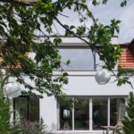 Časť zadnej strany domu s bielou fasádou, viacdielnym presklením na prízemí a panoramatickým oknom na poschodí.