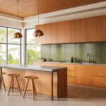 Moderná kuchynská linka v minimalistickom štýle s drevenými skrinkami vo farbe medového dreva, kuchynským ostrovčekom a tmavozelenou zástenou.