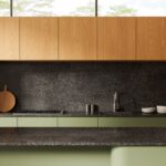 Detail na kuchynskú linku v drevenom dekore v odtieni medového dreva s tmavou zástenou a pracovnou doskou a zelenými dolnými skrinkami.